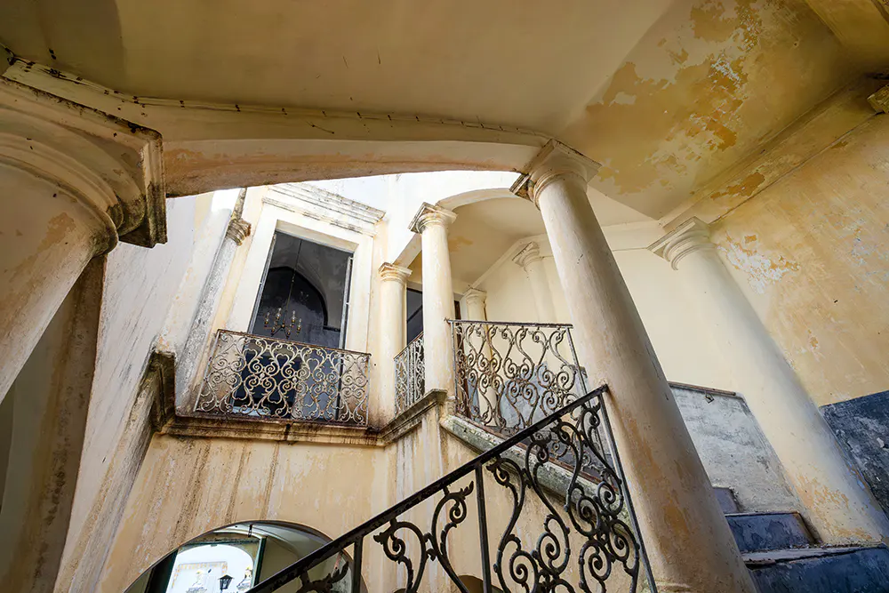 Low-angle shot of a building interior with stairs, columns, and wrought iron railings. The walls are cream-colored with peeling paint.