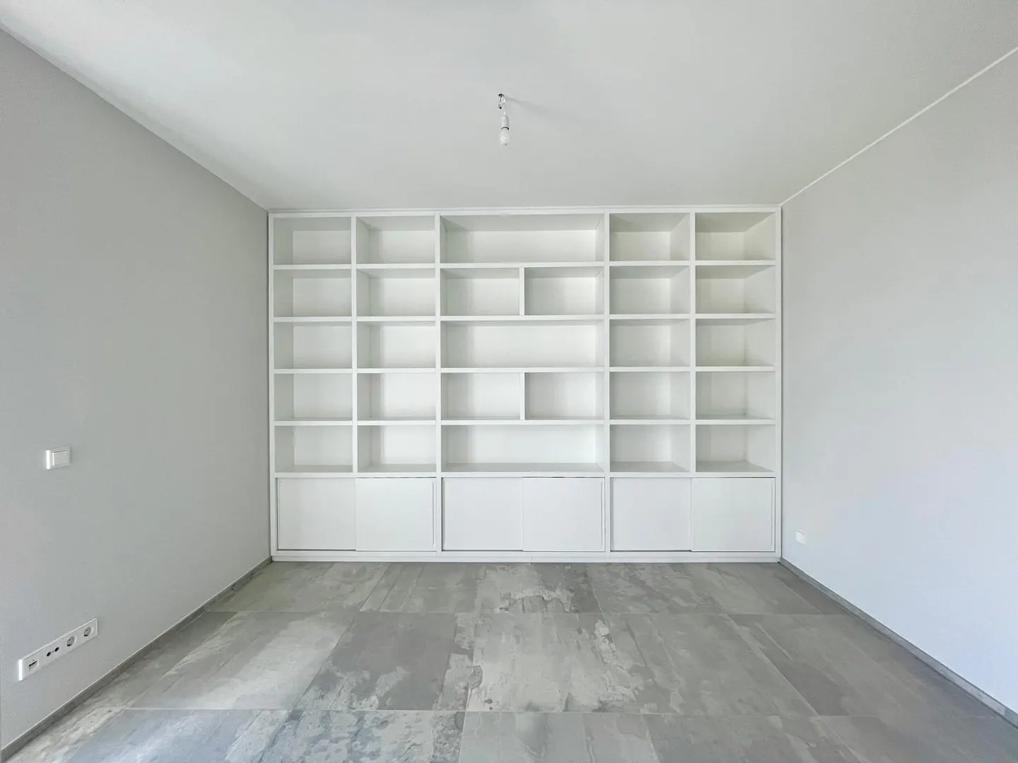 A bright, empty room with gray walls and a gray stone floor features a large, built-in white bookshelf with open shelves and closed cabinets.