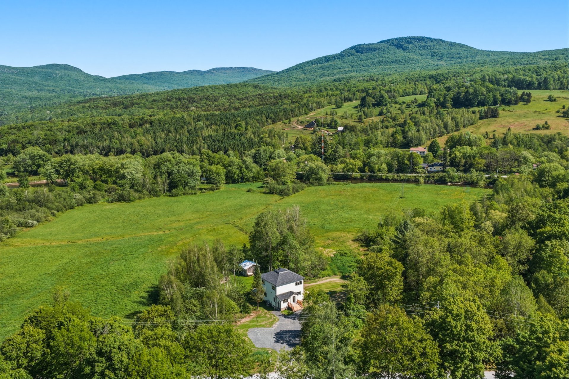 Aerial view of a white house nestled in a lush green valley surrounded by dense forests and rolling mountains under blue sky.