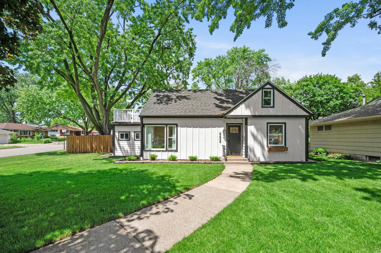 White house with dark trim, a front walkway, and a lush green lawn under large trees on a sunny day.