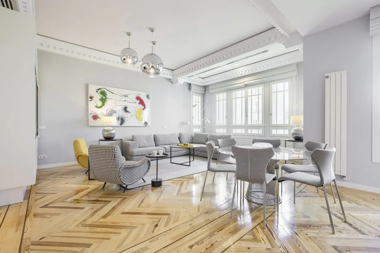Bright, open-concept living and dining area with herringbone wood floors, gray sofa, and modern art. Three chrome pendant lights hang from the ceiling.