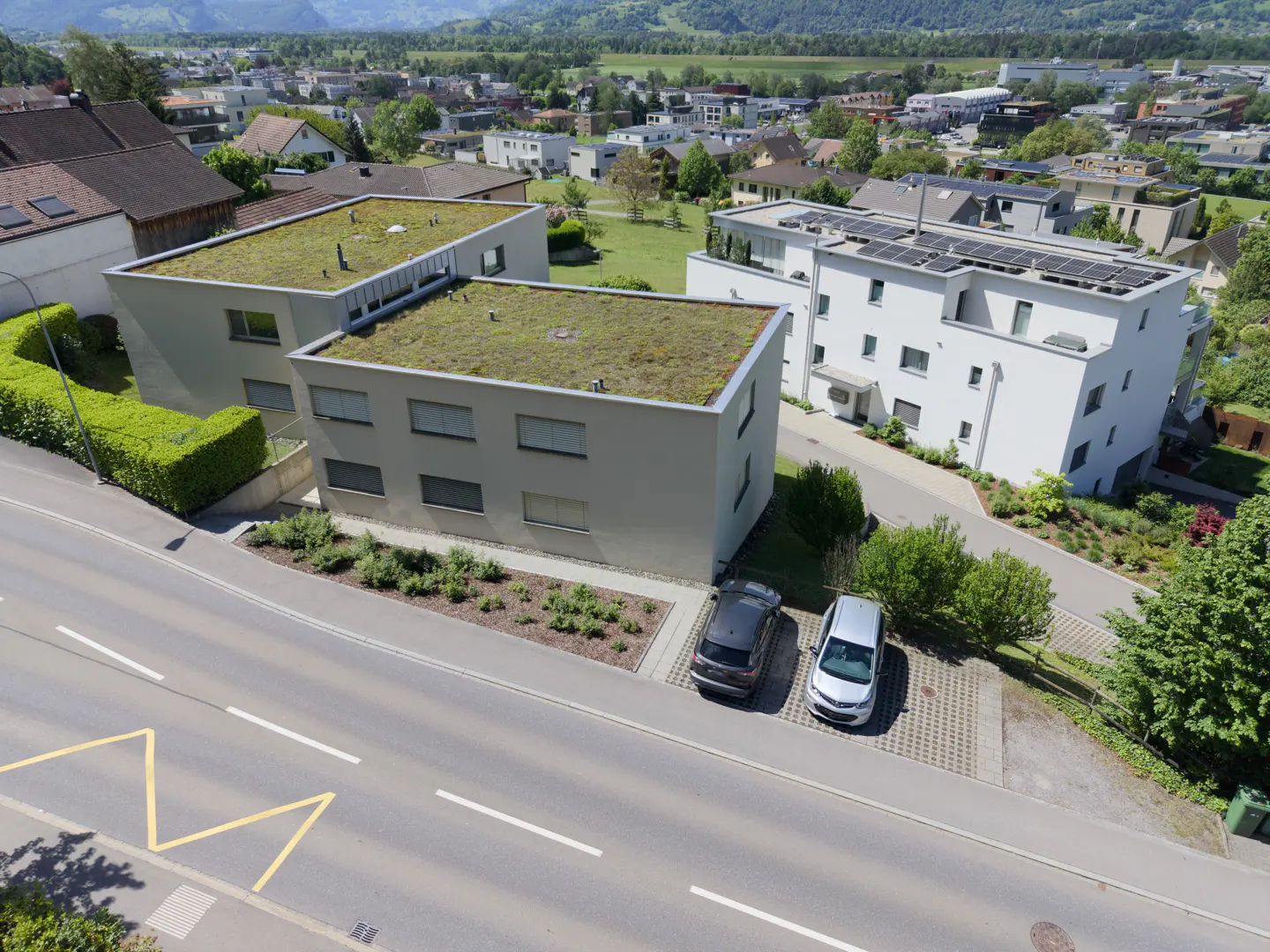 Aerial view of modern, gray buildings with green roofs, parked cars, and mountains in the background.