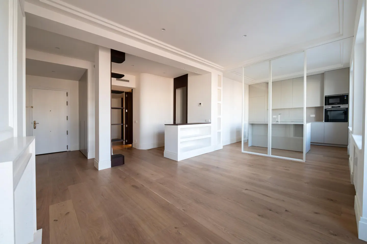 Bright, open apartment with light wood floors. A white kitchen is visible through a glass partition. A white door is on the left.