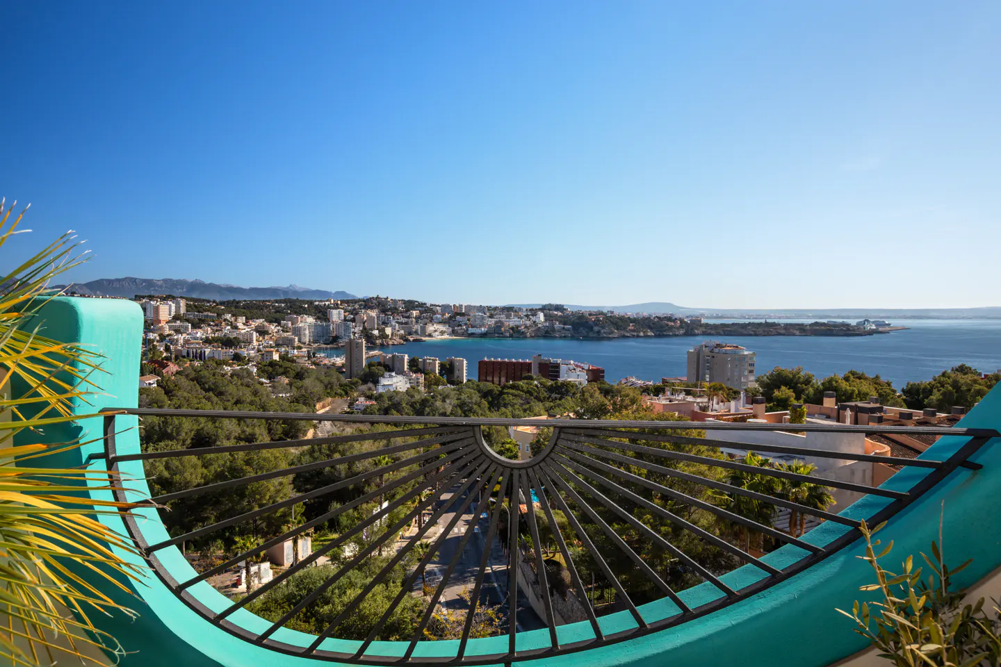 View from a turquoise balcony with black railing overlooking a city, ocean, and blue sky.
