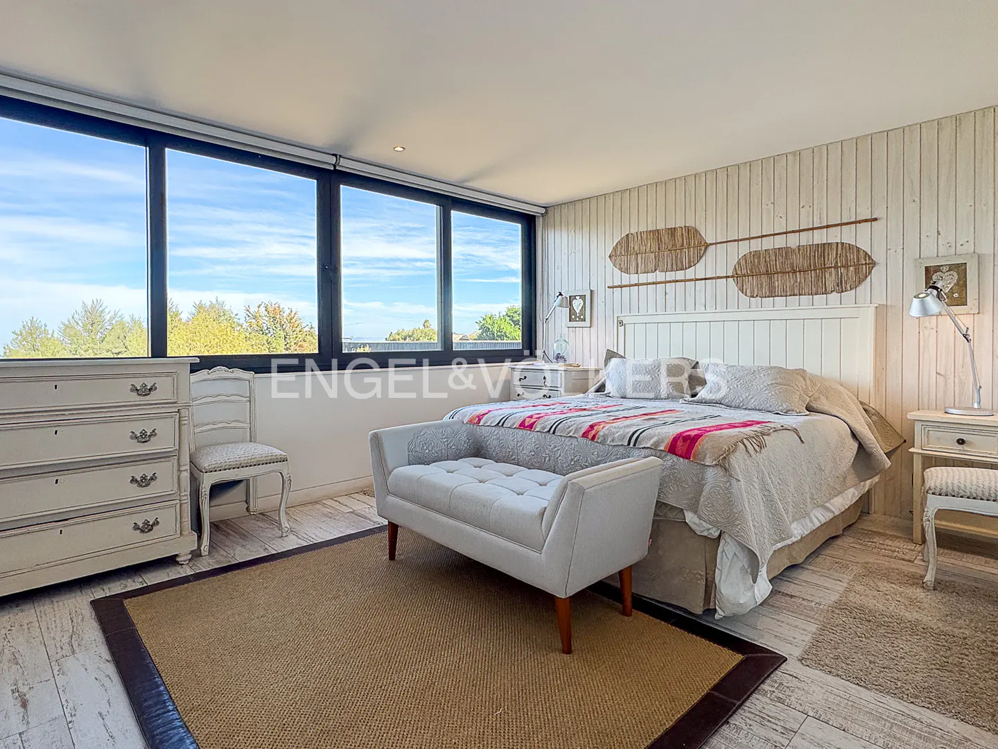 Bedroom with white furniture, a large bed with patterned bedding, and a bench in front of a window with a view of trees and sky.