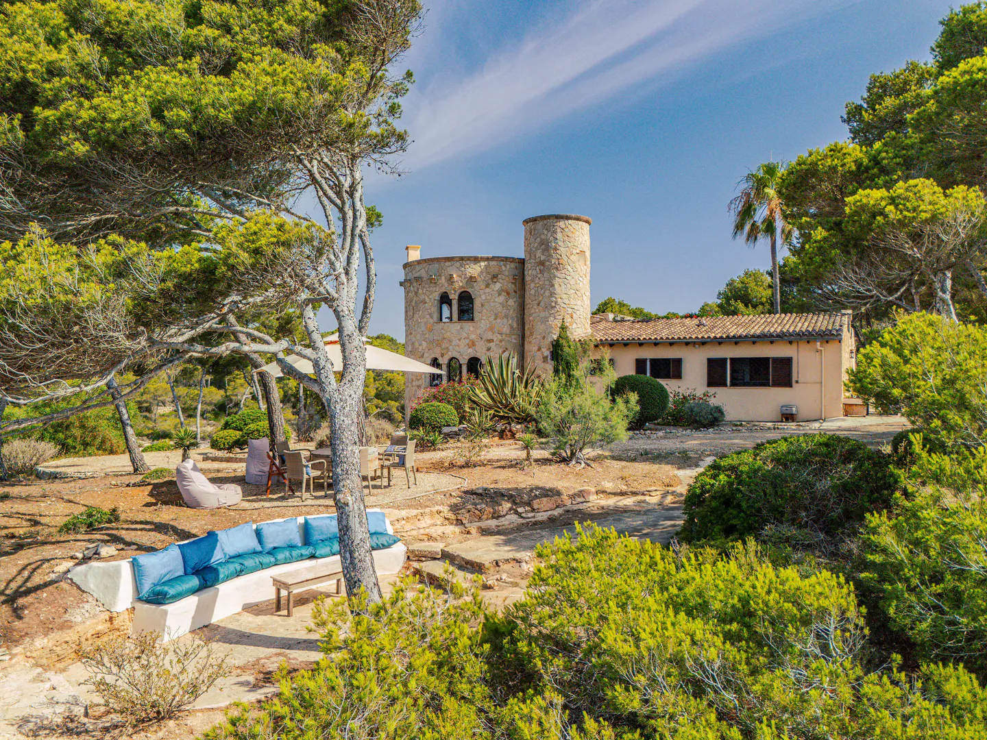Exterior view of a stone house with a tower, surrounded by trees and a blue cushioned bench in the foreground.
