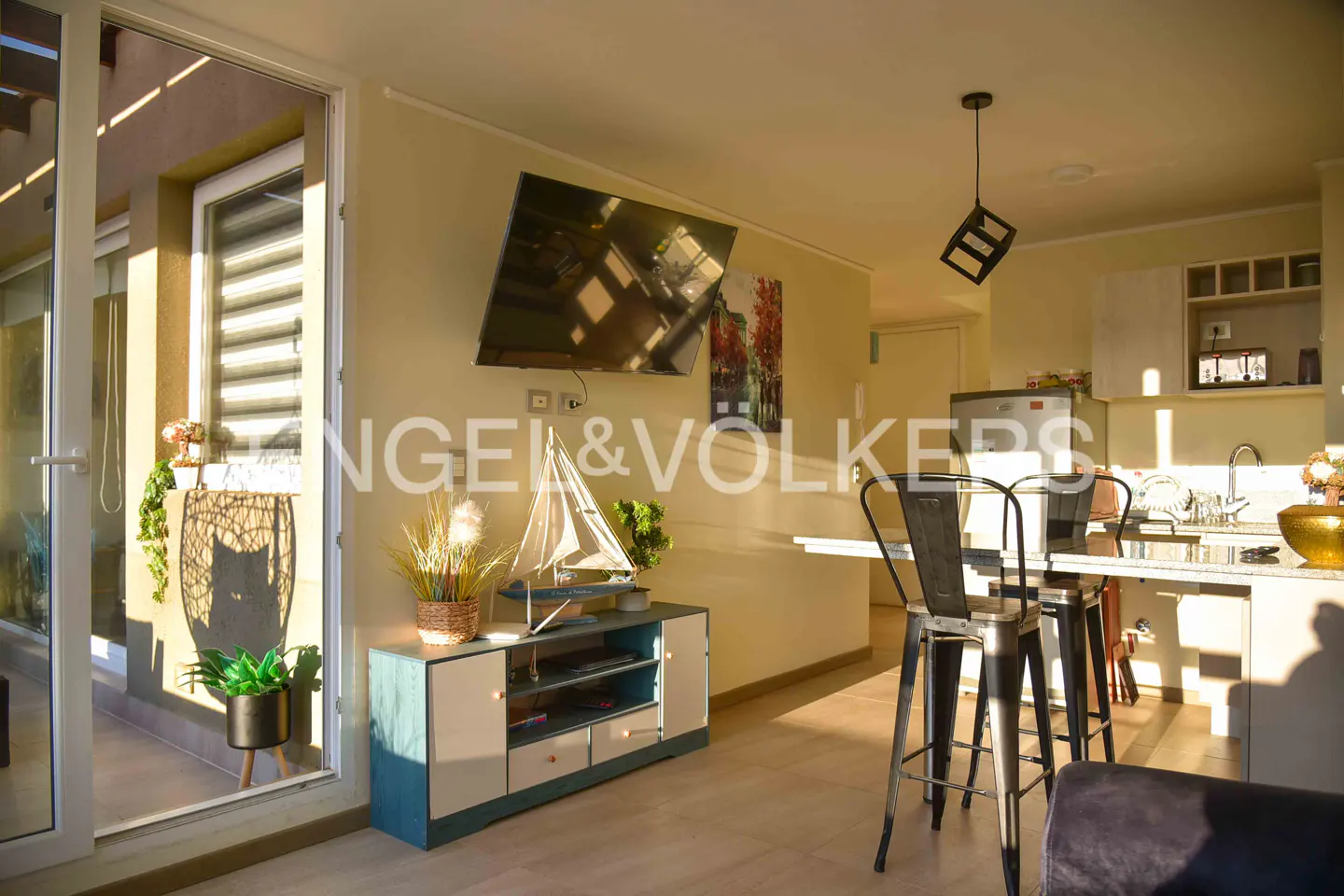 Bright living room with TV, blue cabinet, and bar stools at a kitchen counter. Balcony visible through sliding glass door.