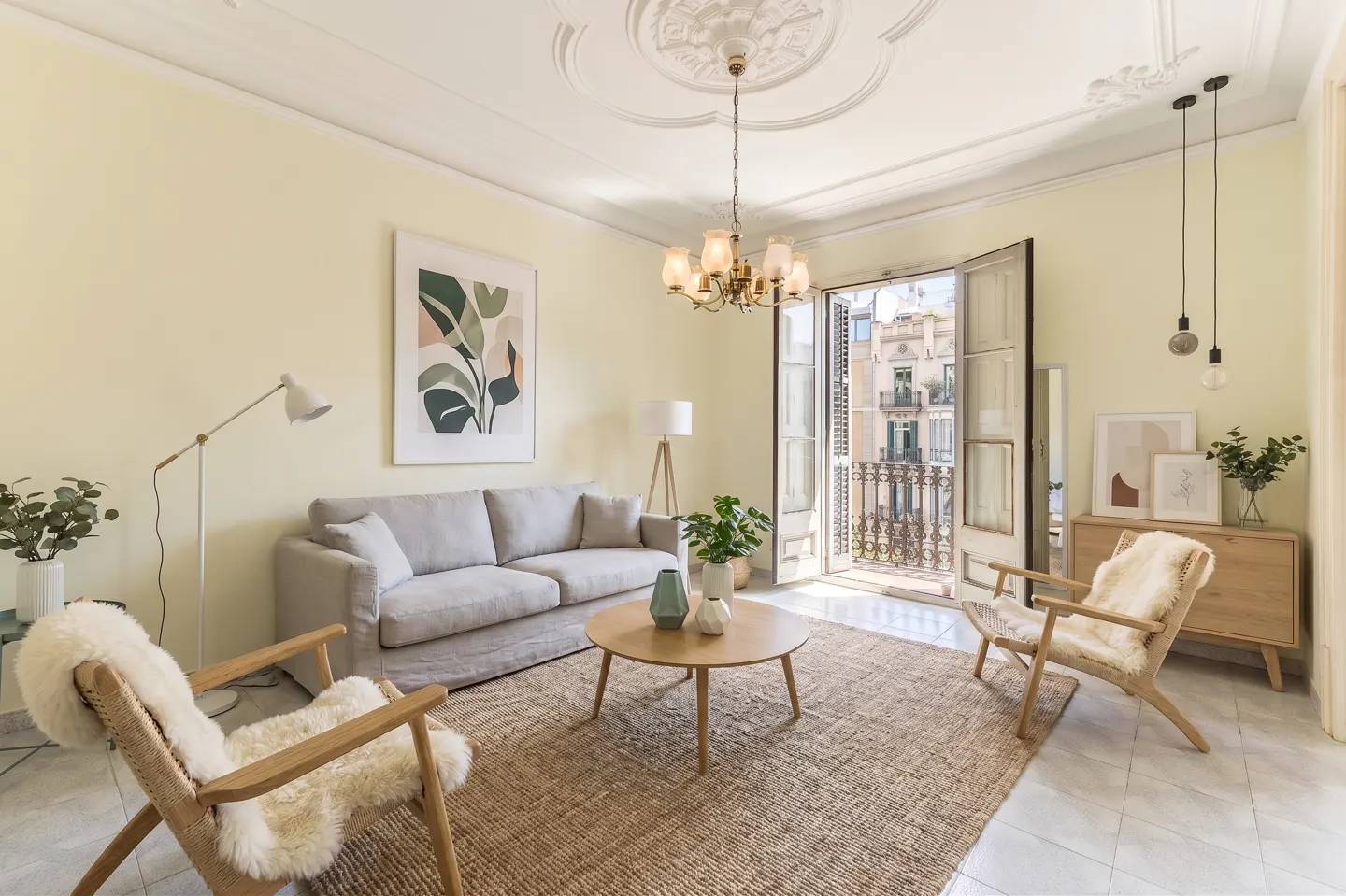 Bright living room with a gray sofa, round table, and two chairs on a jute rug. Balcony doors open to a city view.