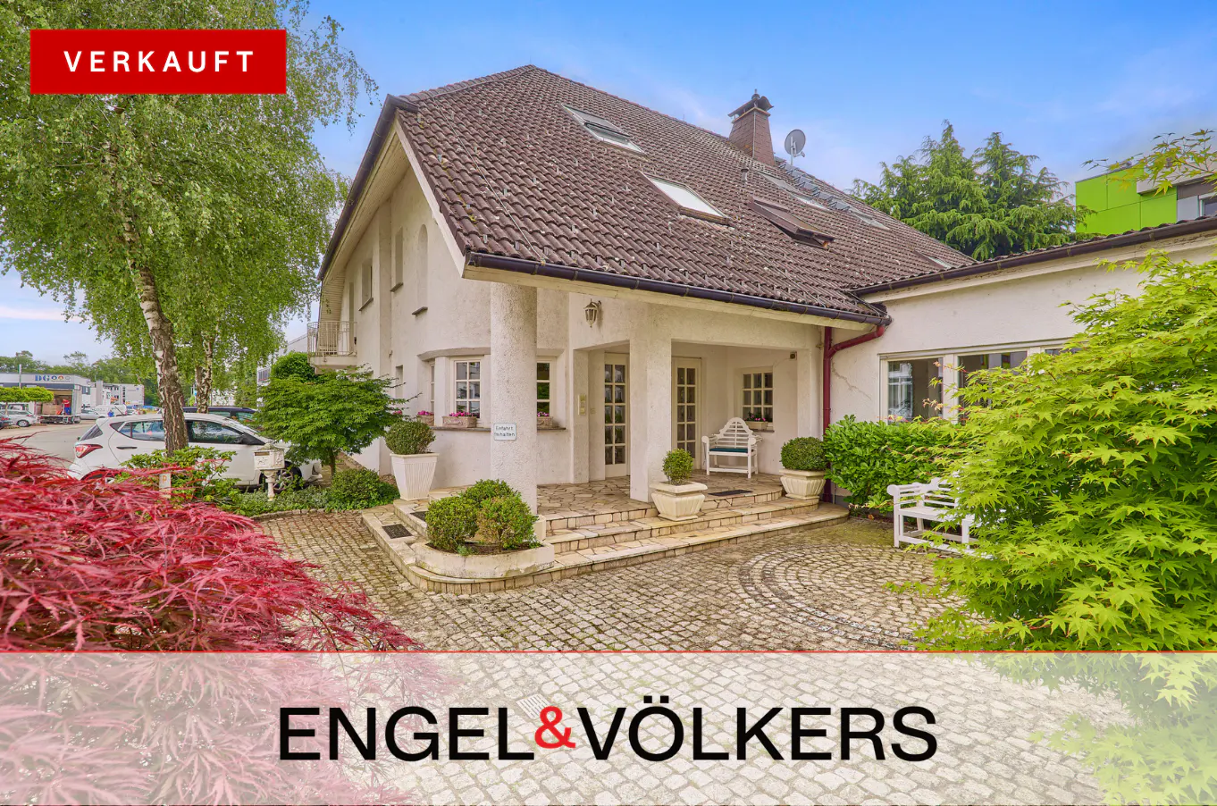 Exterior of a two-story white house with a brown roof, a stone walkway, and green foliage. "Verkauft" banner in the upper left corner.