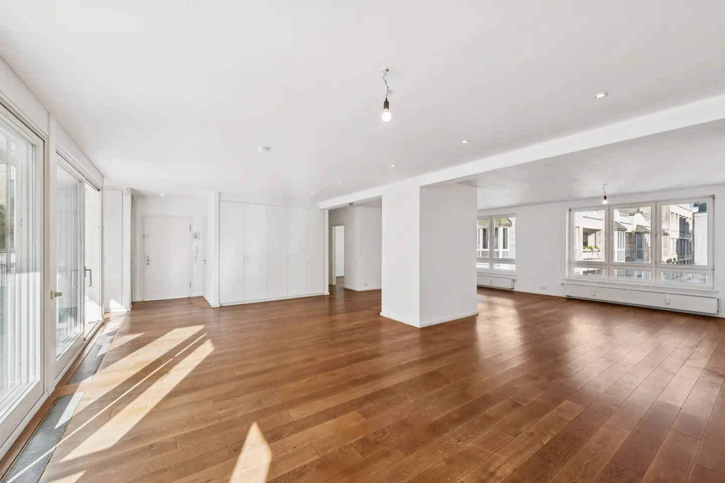 Bright, empty room with hardwood floors, white walls, and large windows. Sunlight streams in, highlighting the spacious interior.