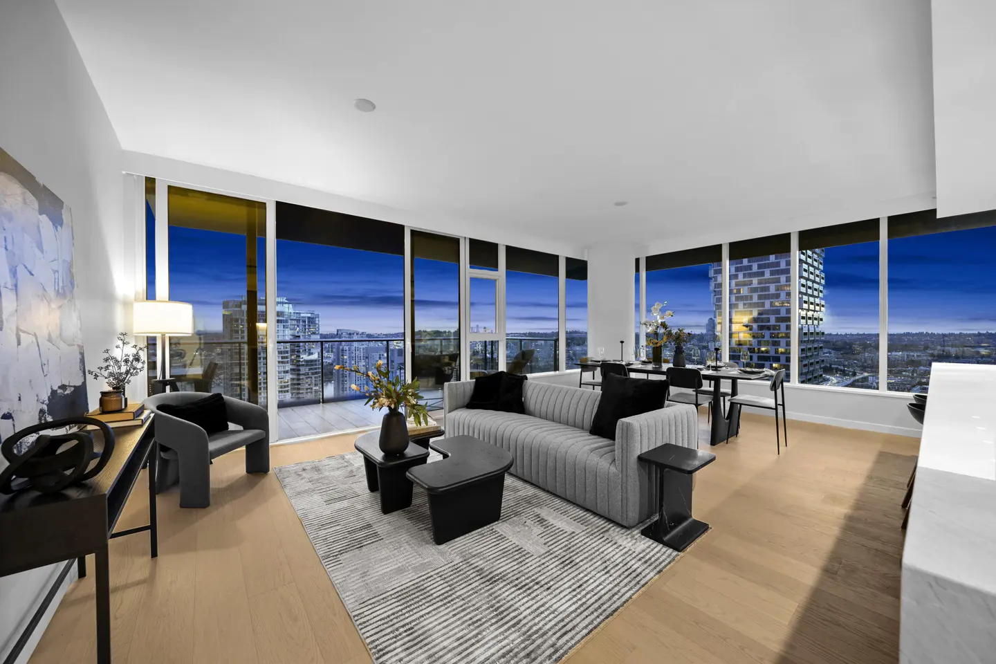Bright, modern condo living room with floor-to-ceiling windows overlooking a city skyline at dusk. Gray sofa, black accents, and light wood floors.