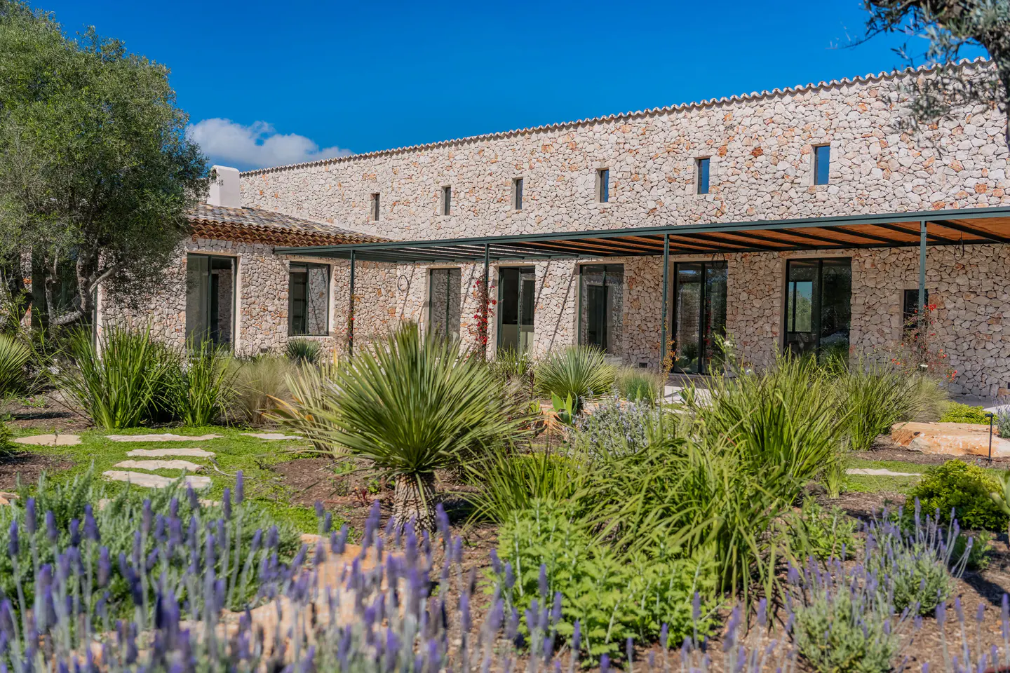 Stone house with black framed windows and a covered patio. Lavender and green plants in the foreground. Blue sky.