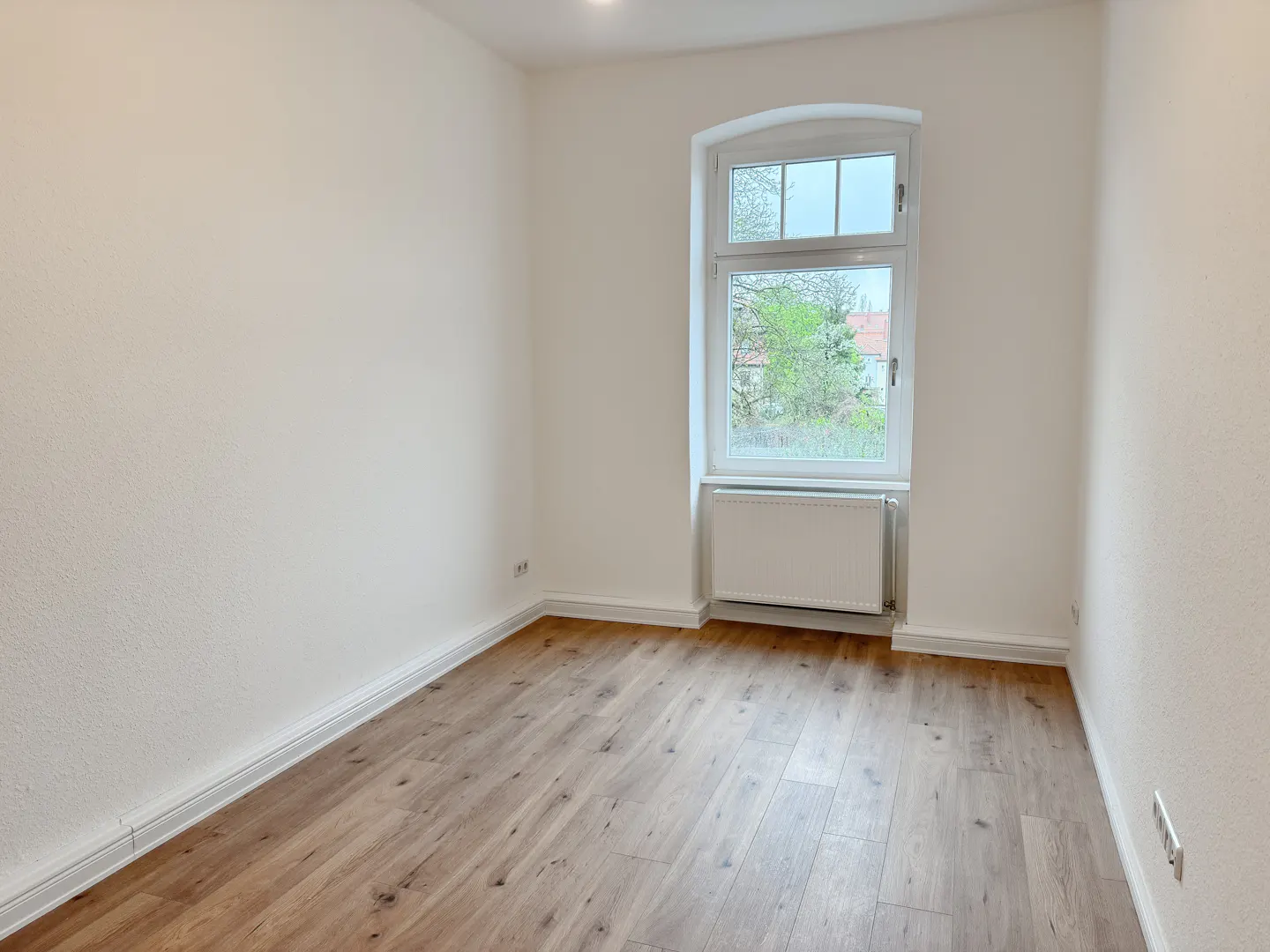 Bright, empty room with light wood floors, white walls, and a white-framed window showing green trees outside.