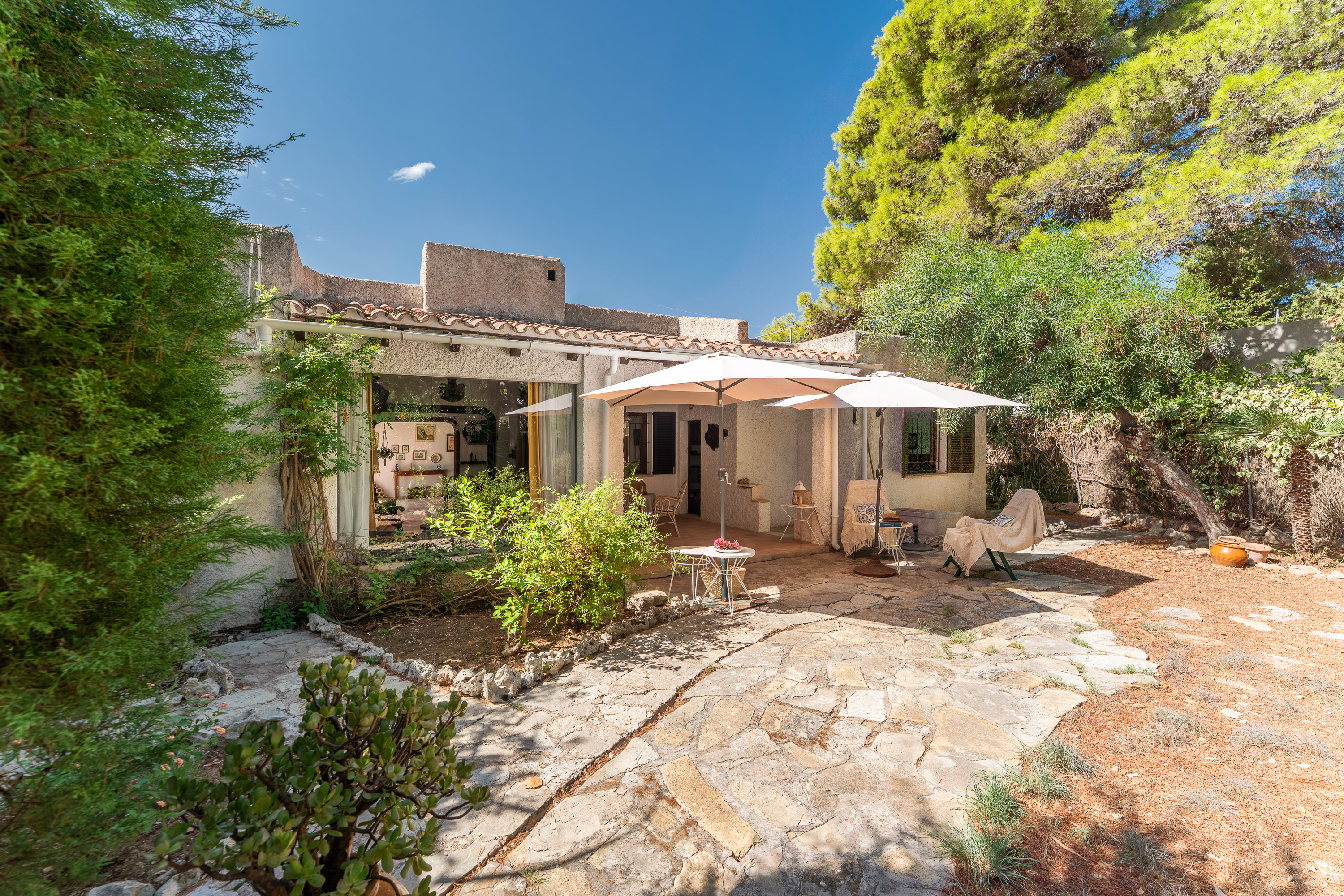 Stone patio of a Mediterranean-style home with white umbrellas, outdoor seating, and lush trees under a clear blue sky. [AI-generated text]