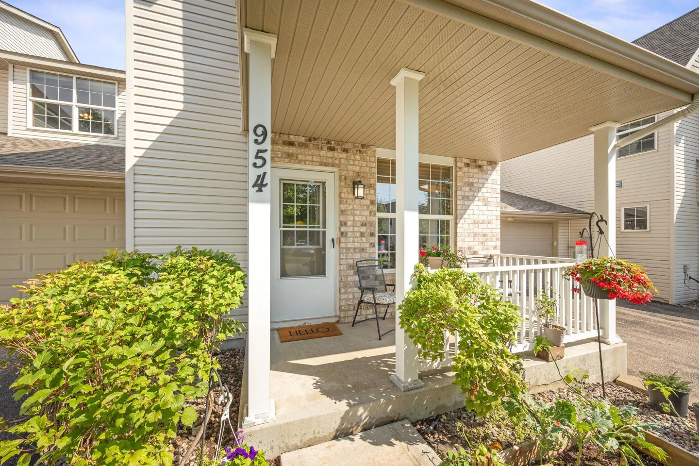 Exterior view of a two-story townhouse with a covered porch, white door, and the address "954" displayed. Green bushes and hanging flower baskets add color.