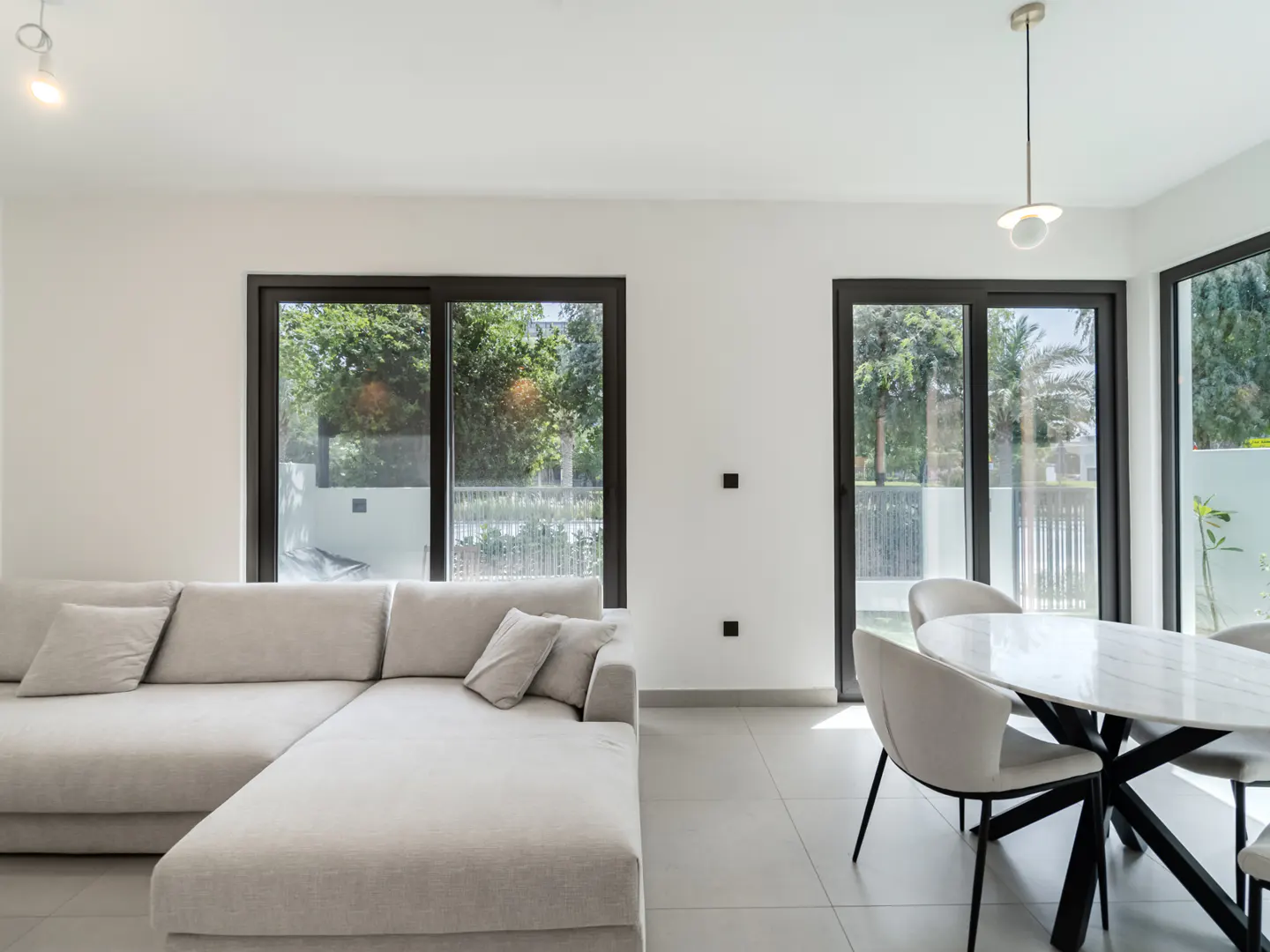 Bright, modern living room with a beige sectional sofa, marble dining table, and black-framed windows overlooking lush greenery.