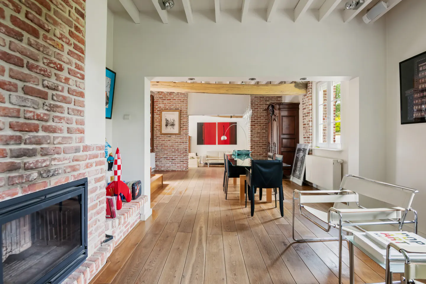 A bright, open-concept living and dining area with hardwood floors, a brick fireplace, and modern furniture.