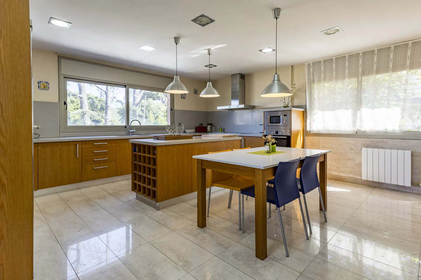 Modern kitchen with light wood cabinets, a central island, white dining table, blue chairs, and large windows.