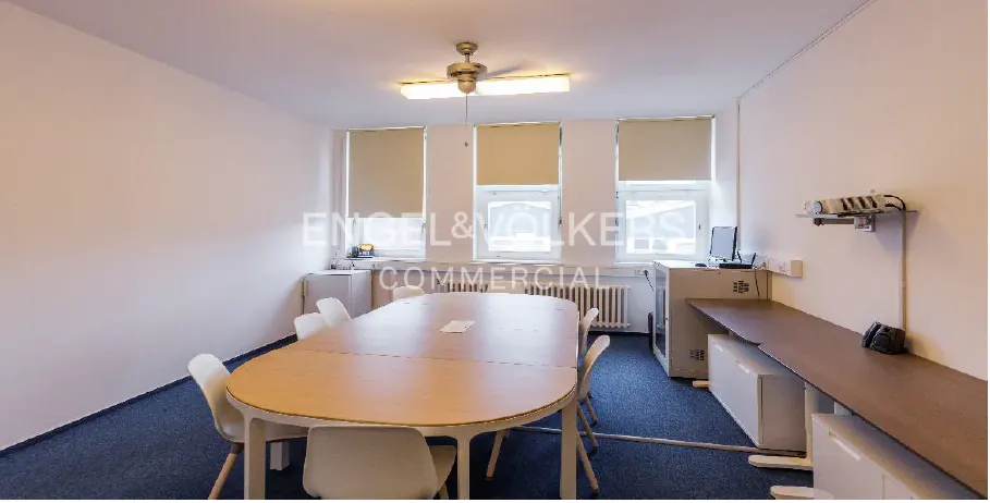 A bright office meeting room with a large oval wooden table, white chairs, blue carpet, and three windows with beige blinds.