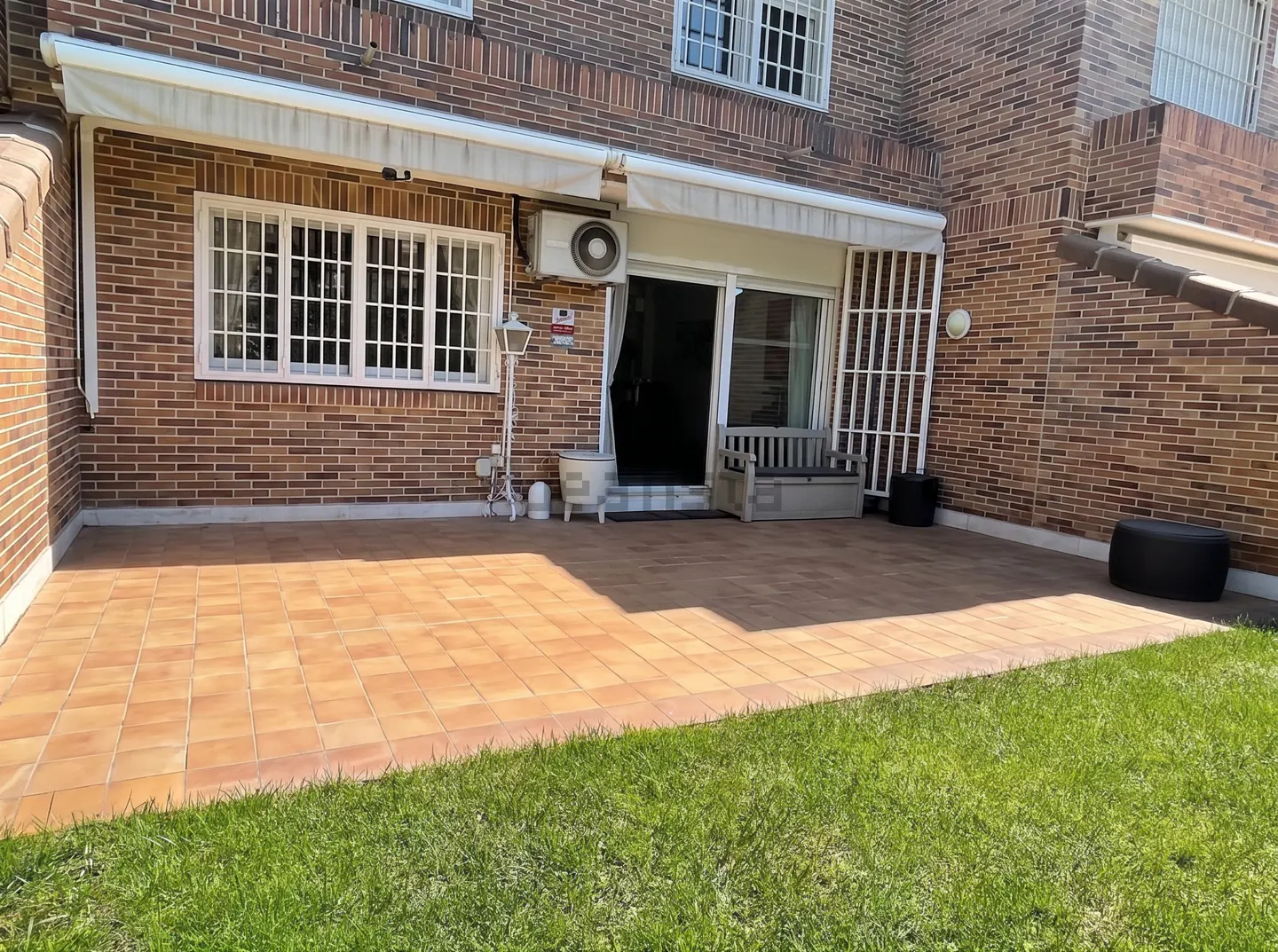 Brick house with a tiled patio and green lawn. A white awning covers the sliding glass door and window with white bars.