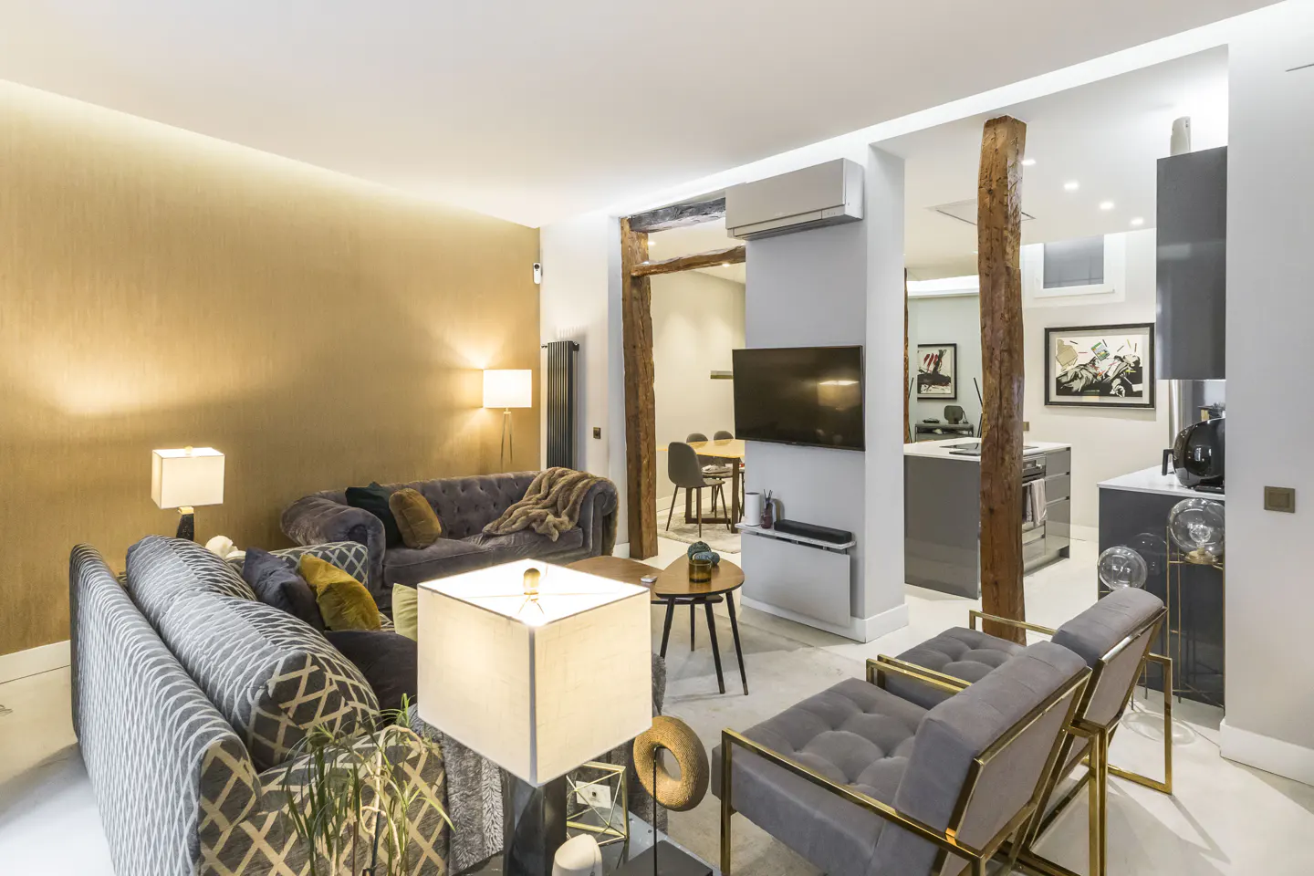 A modern living room with gray sofas, gold accents, and a view into a gray kitchen. A TV is mounted on the wall.