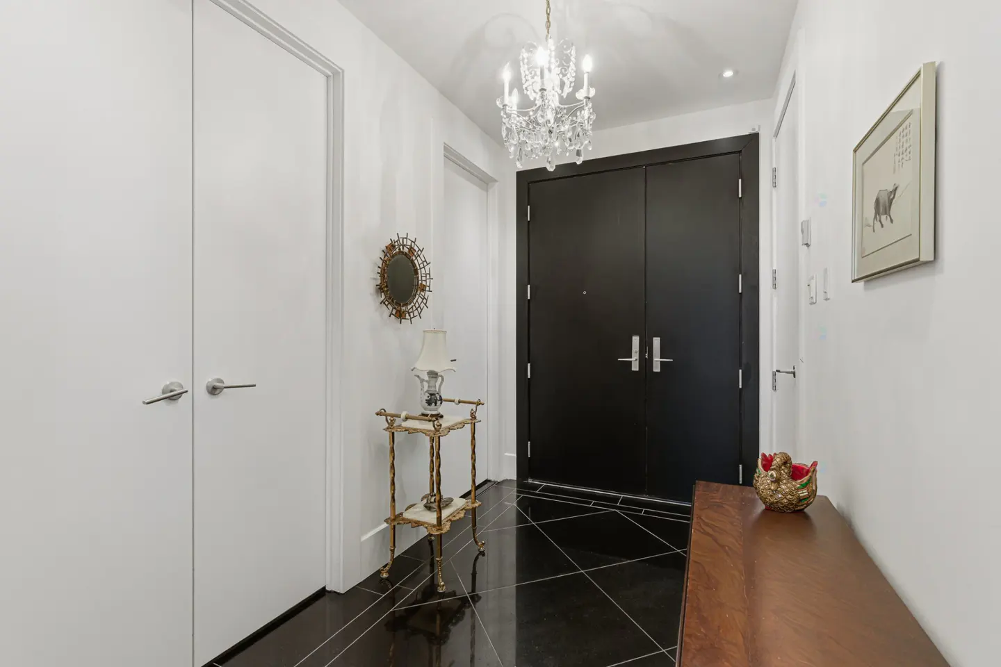 A foyer with black tile floors, white walls, and a black double door. A crystal chandelier hangs from the ceiling.