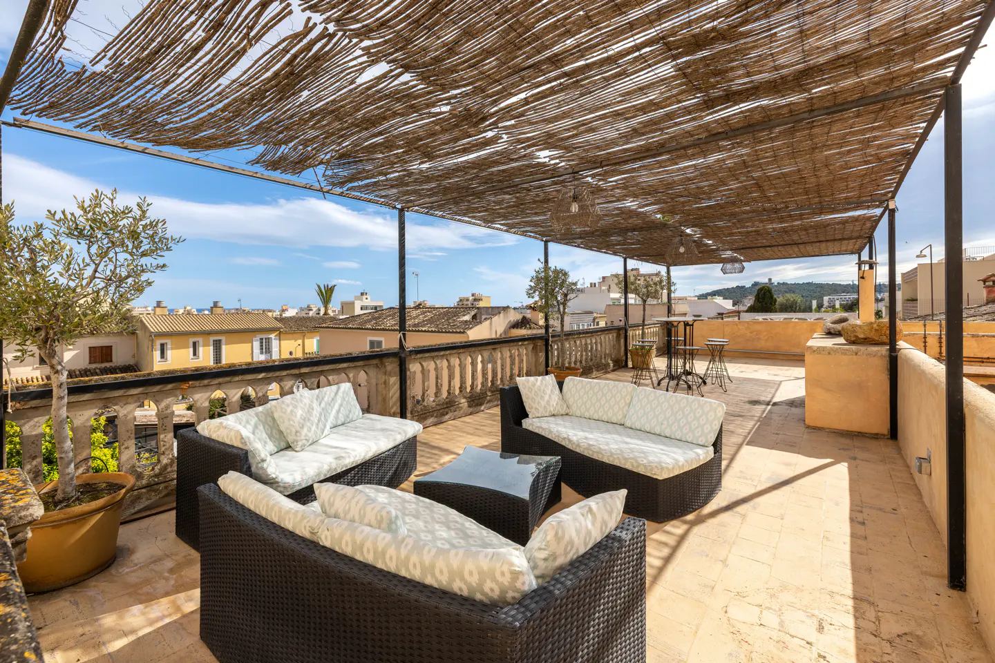 Rooftop patio with wicker furniture, a glass table, and a straw awning. The patio overlooks a town with yellow buildings and a blue sky.