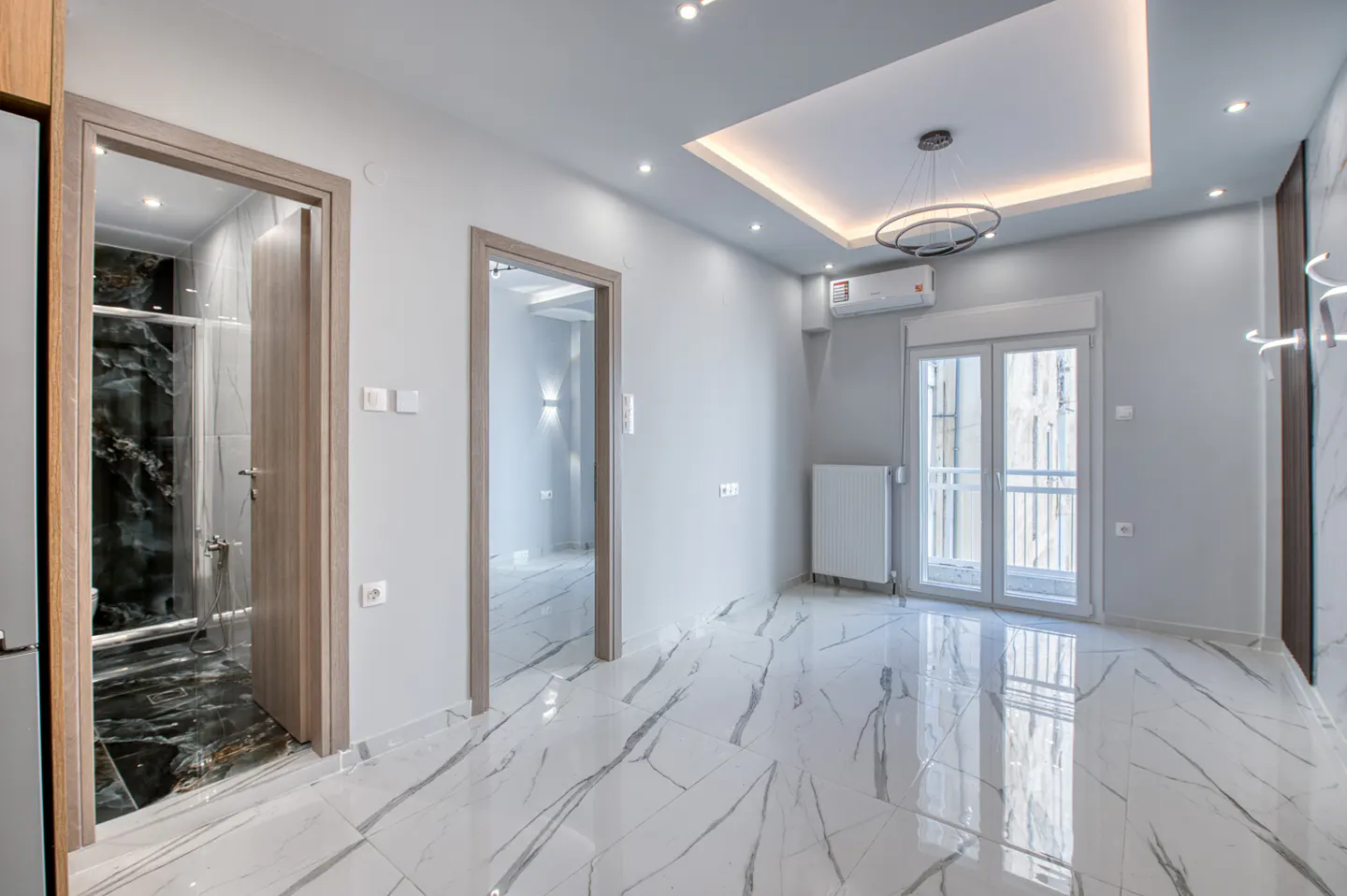 Bright, empty room with white marble floors, gray walls, and two open doorways. One doorway leads to a bathroom with black marble walls.