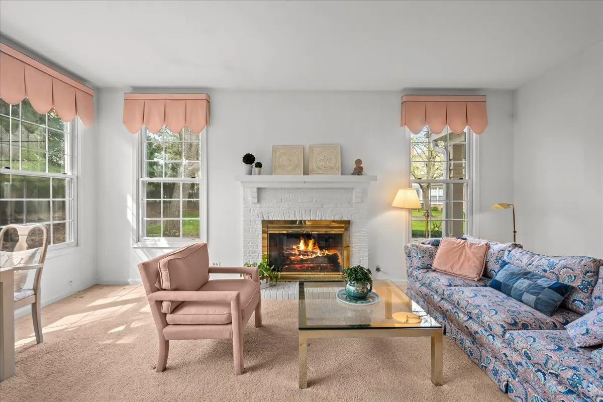 Bright living room with white walls, pink armchair, floral sofa, and a fireplace with a white mantel. Large windows with pink scalloped valances.