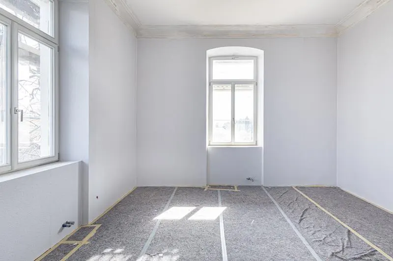 Bright, empty room with white walls, two windows, and gray floor covering with tape lines. Sunlight shines on the floor.