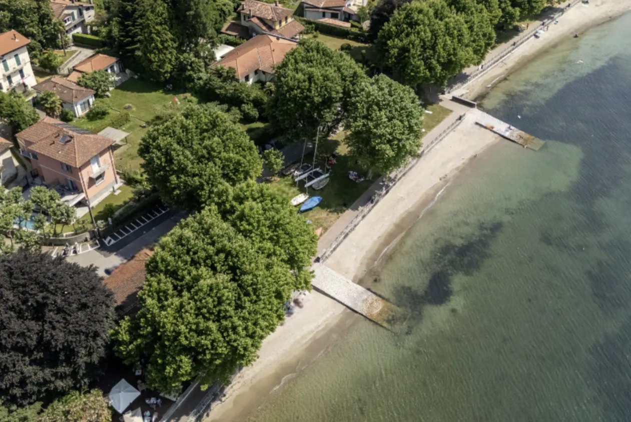 Aerial view of a sandy beach with green trees, houses, and clear water. A stone pier extends into the water. Boats are on the shore.