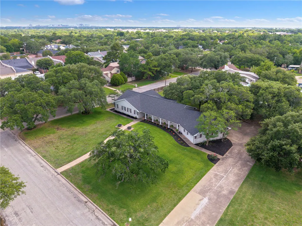 Aerial view of a white ranch-style home with a dark roof, large front lawn, mature trees, and a concrete driveway.