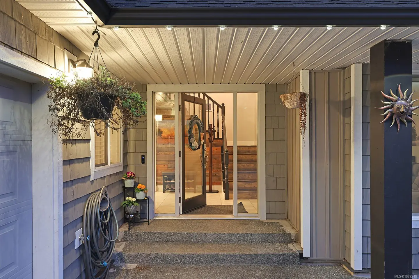 Exterior view of a home's front entrance with an open door, stairs, and hanging plants. The house has gray siding and a sun decoration on a pillar.
