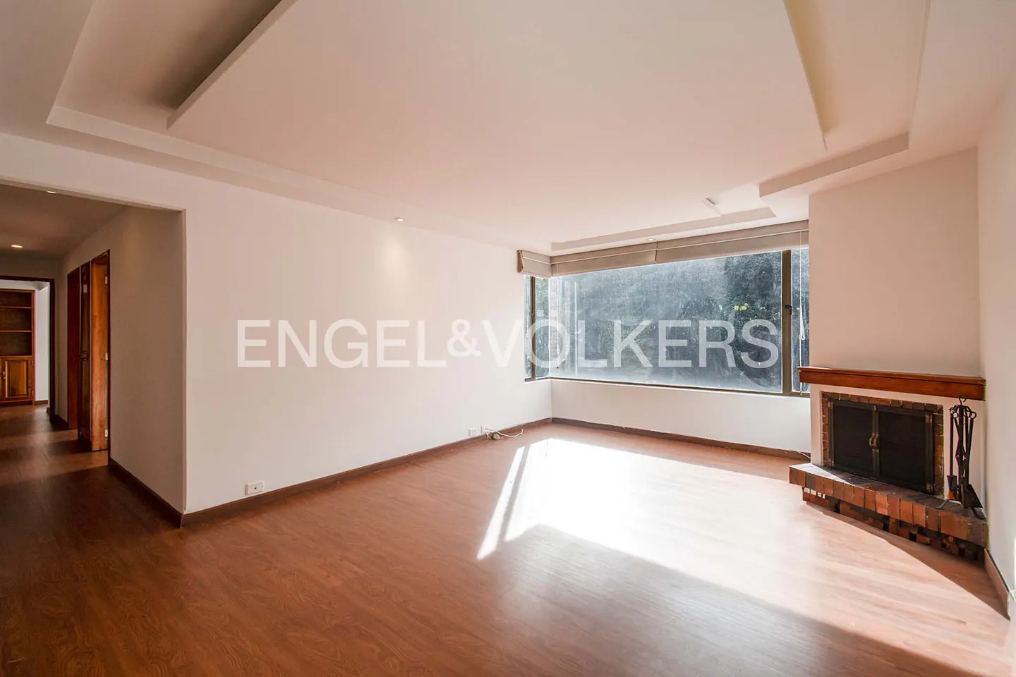 Bright, empty living room with hardwood floors, white walls, and a brick fireplace. A large window offers natural light. Engel & Volkers logo on the wall.