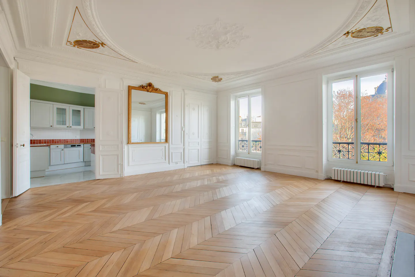 Bright, empty room with herringbone wood floors, white walls, and large windows. A gold-framed mirror hangs on the wall, and a kitchen is visible through a doorway.