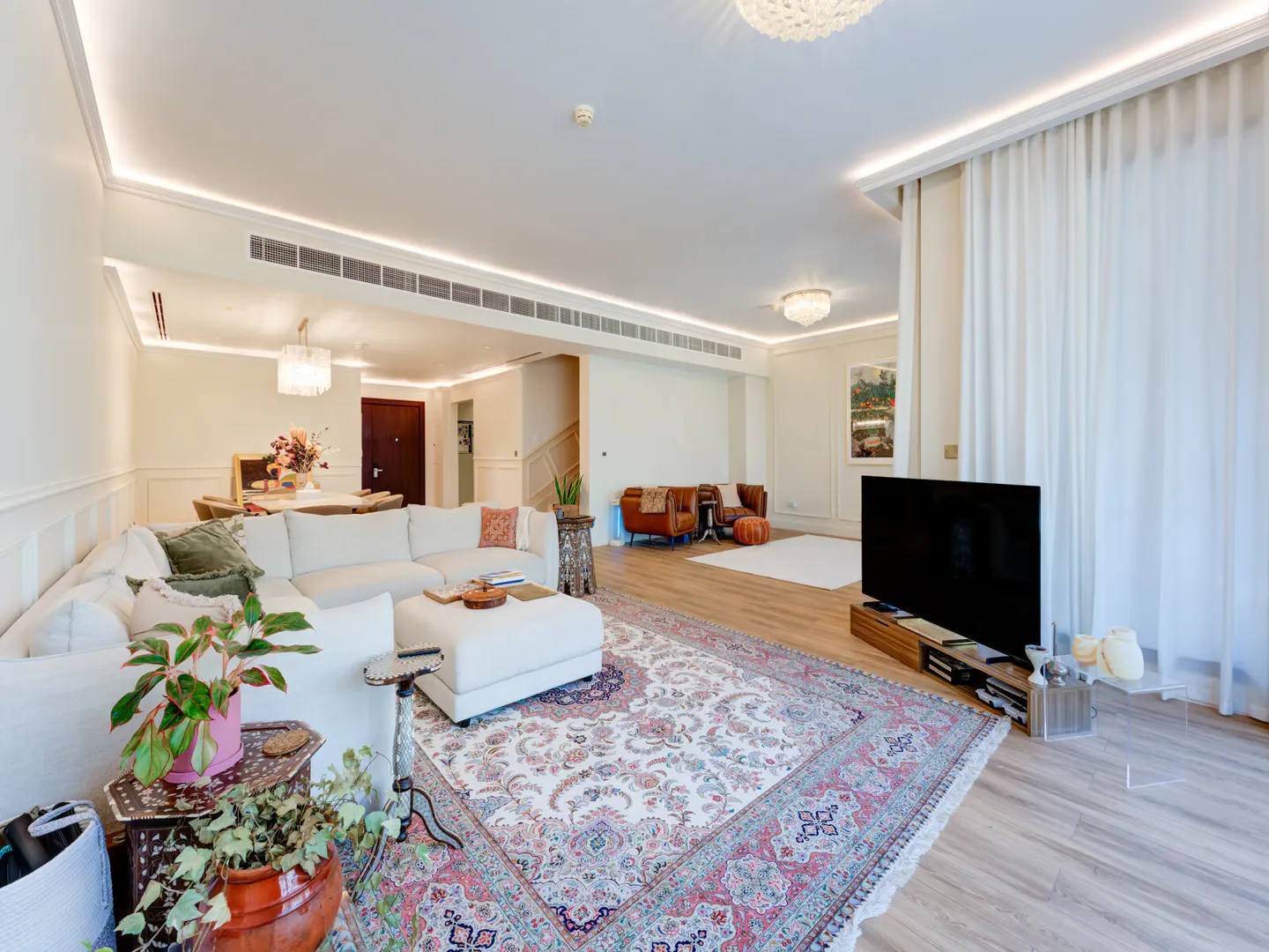 Bright, open living room with a white sectional sofa, patterned rug, and a large TV. Dining area and brown leather chairs are visible in the background.