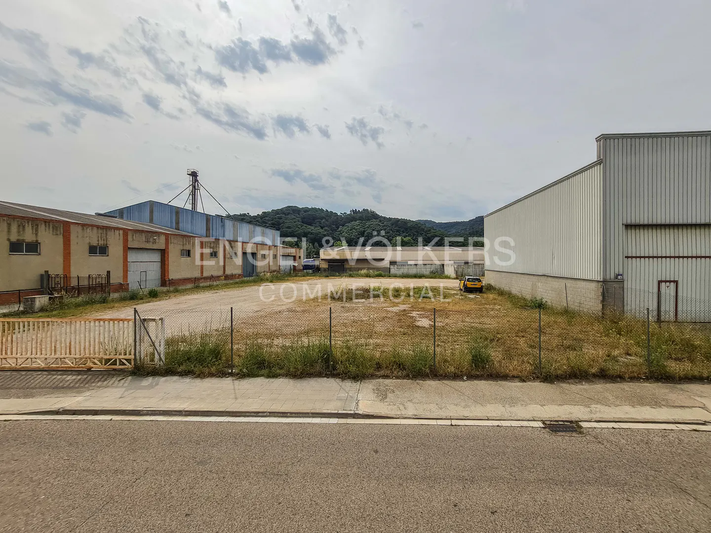 An empty dirt lot sits between two industrial warehouse buildings under a cloudy sky, viewed from a paved street.
