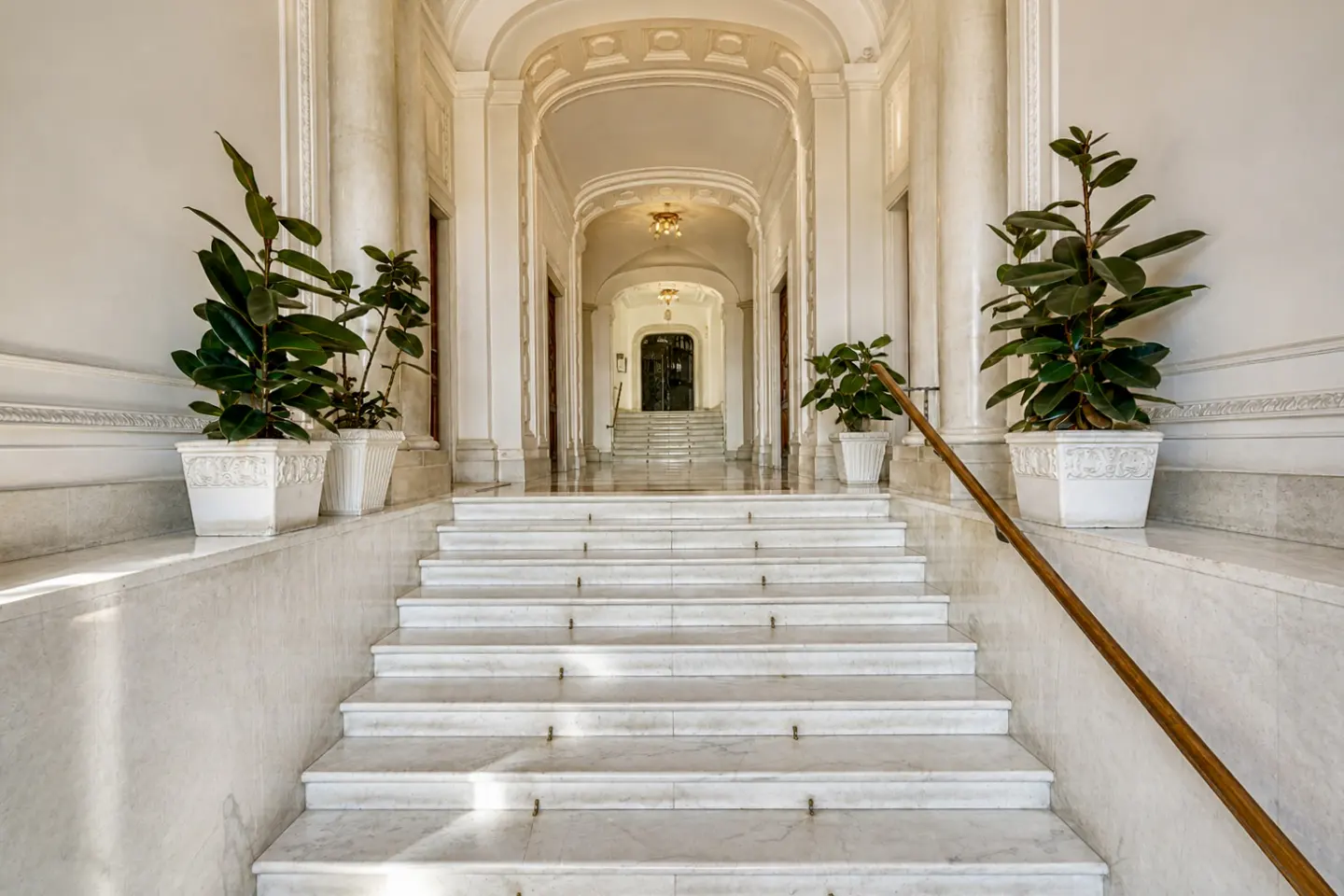 Elegant marble staircase with wood handrail, leading to arched hallway with potted plants.