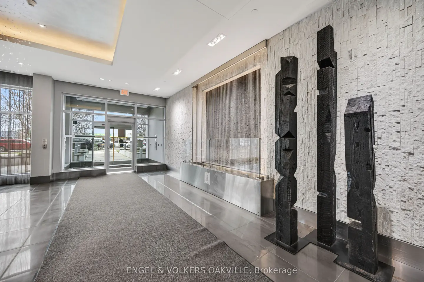 Lobby with gray carpet, glass doors, stone wall, and three dark wood sculptures. A water feature is in the background.