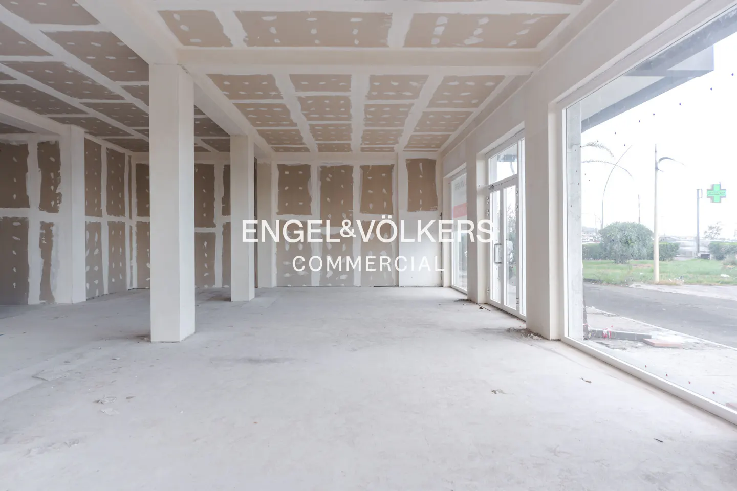 Unfinished commercial space with white support columns, drywall walls and ceiling, and large windows overlooking a street.