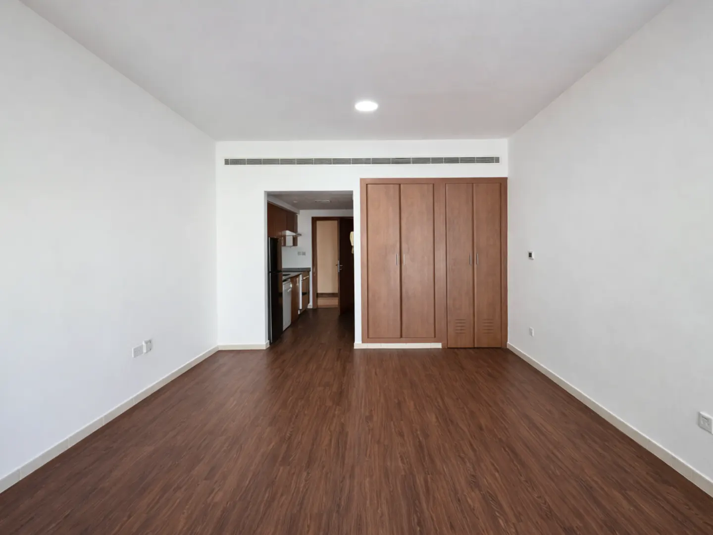 Empty room with white walls, wood-look flooring, built-in wooden cabinets, and a view into a small kitchen.