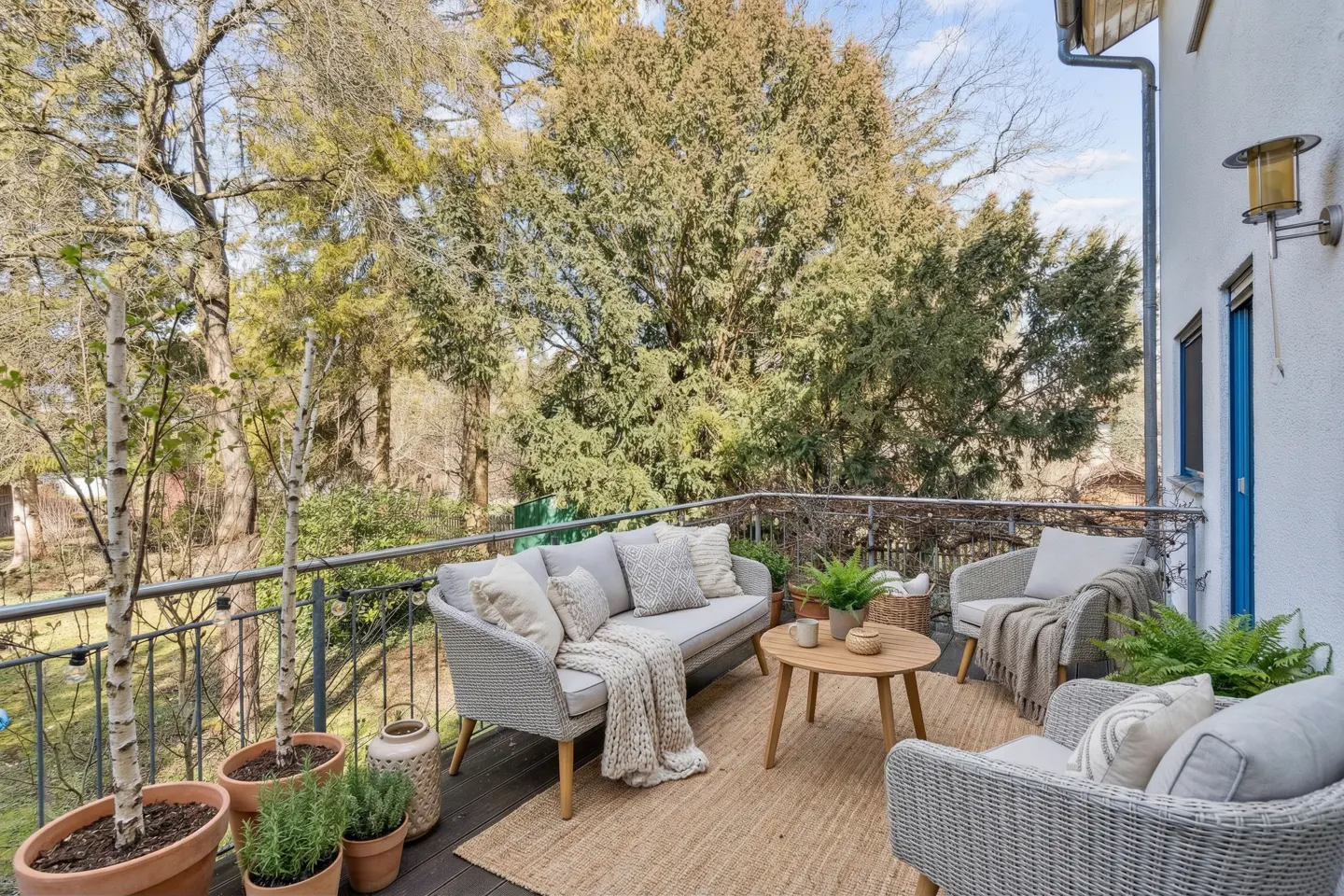 Balcony with gray wicker furniture, including a sofa, chairs, and a round wooden table on a jute rug. Potted plants and trees surround the space.