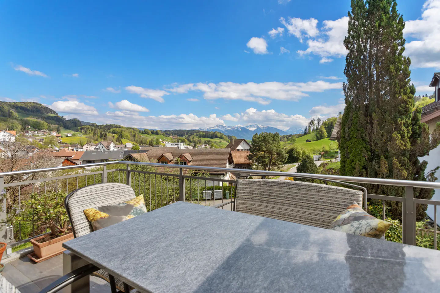 Balcony view with table, chairs, and mountain vista. Blue sky with clouds above green hills and houses.