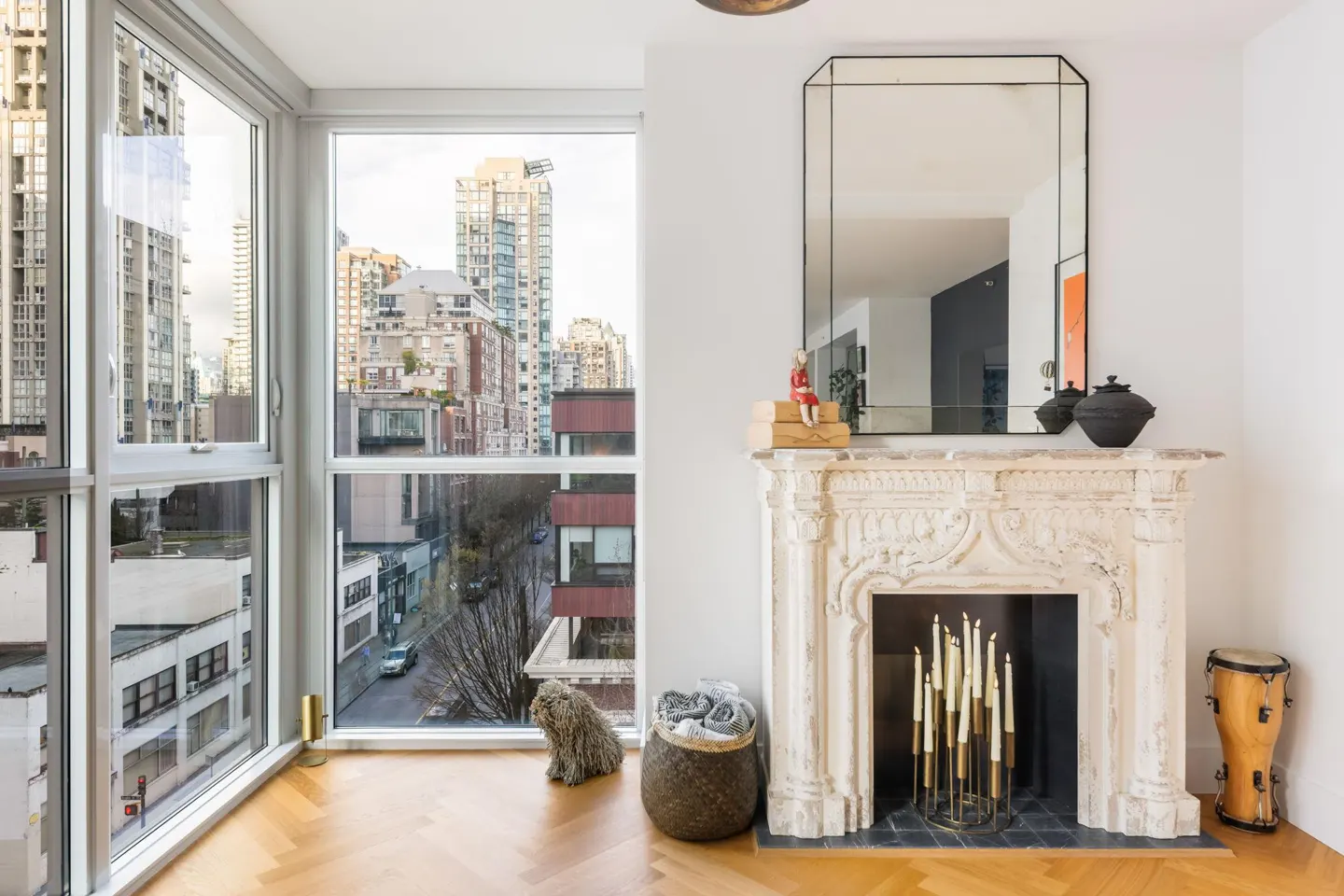 Bright living room with a large window overlooking a city. A white fireplace with a mirror above it is on the right. A drum is next to the fireplace.