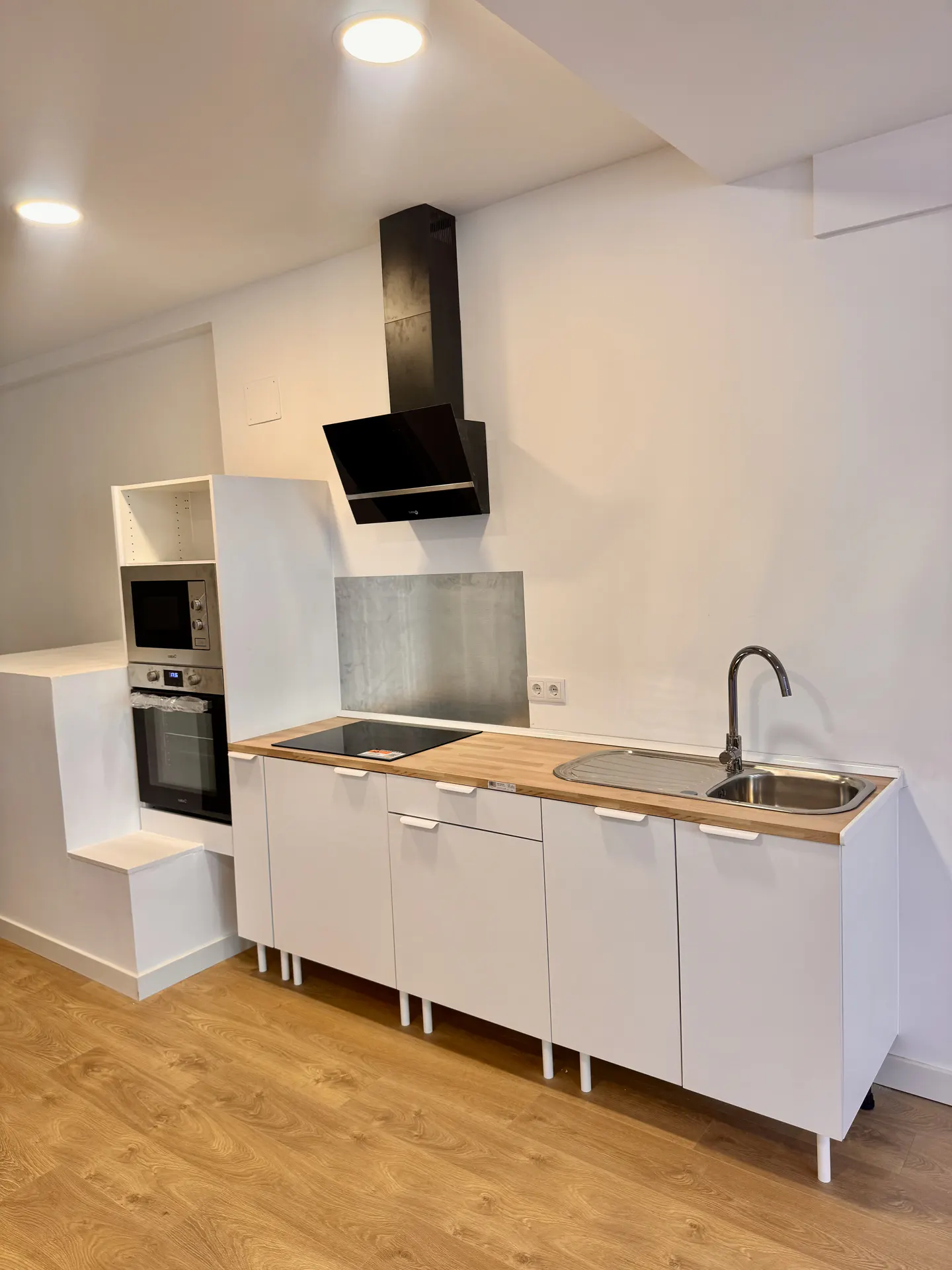A modern kitchen with white cabinets, wood countertops, and stainless steel appliances. A black range hood is mounted above the stove. The floor is light brown wood.