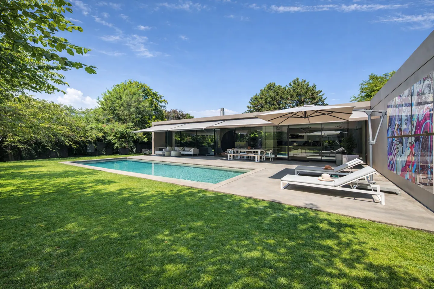 Modern home exterior with a pool, patio furniture, and a green lawn under a blue sky.