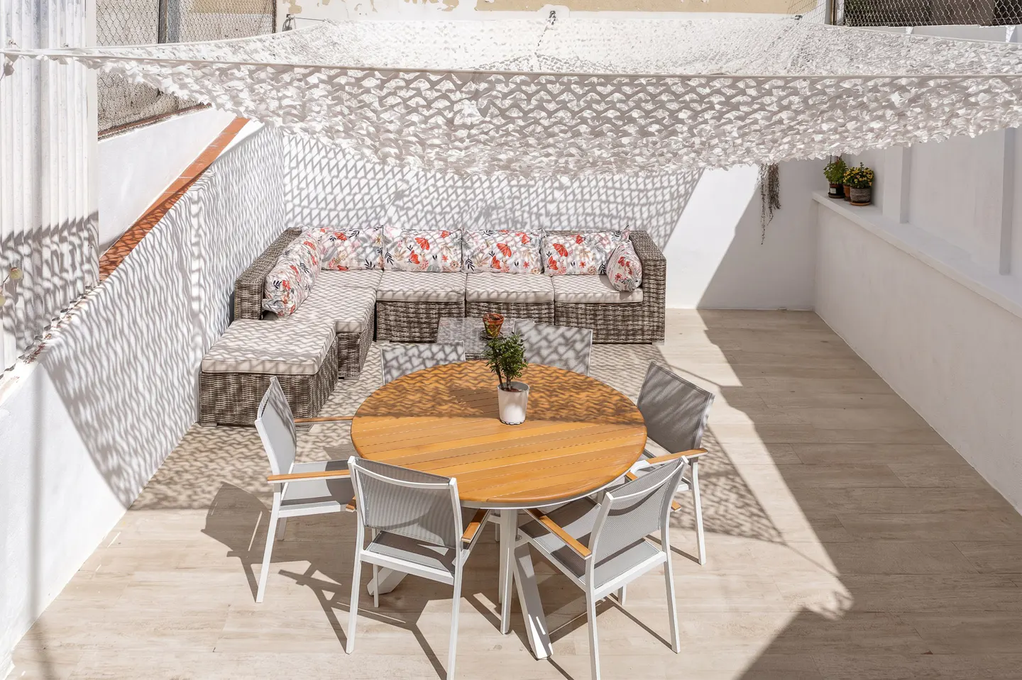 Outdoor patio with a round wooden table, six chairs, and a wicker sectional sofa with floral pillows. A white shade sail provides cover.