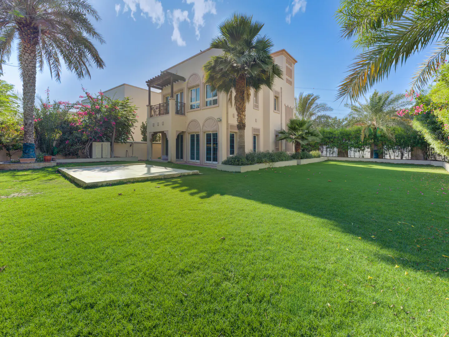 Two-story beige house with a large green lawn, palm trees, and a blue sky.