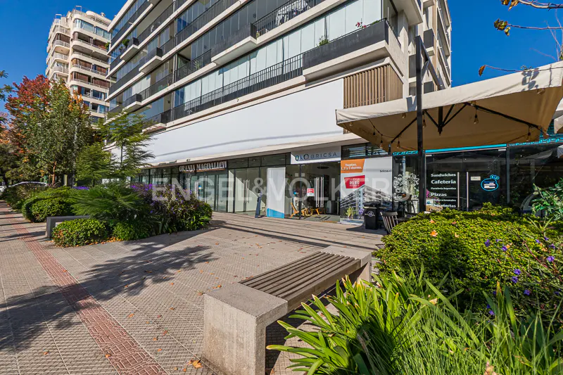 Street view of an Engel & Völkers real estate office in a modern building with shops, benches, and greenery on a sunny day.
