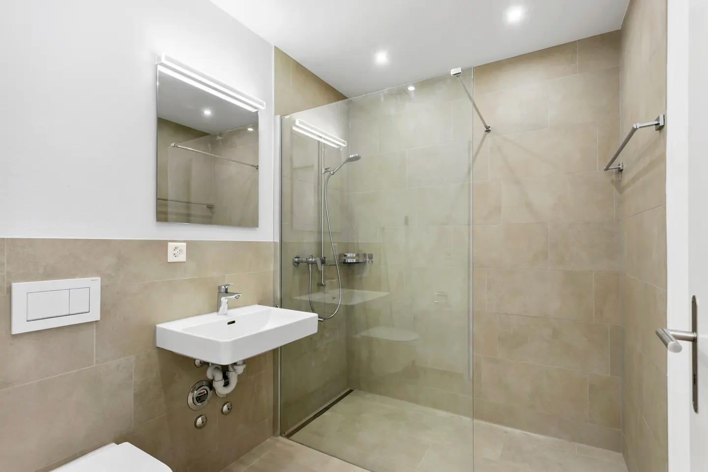 Modern bathroom with beige tile, a glass-enclosed shower, a white sink, and a mirror with a light fixture.